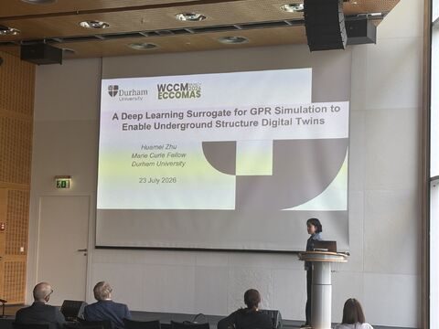 Huamei Zhu giving a talk at WCCM-ECCOMAS 2026 in Munich, with the title slide on screen.
